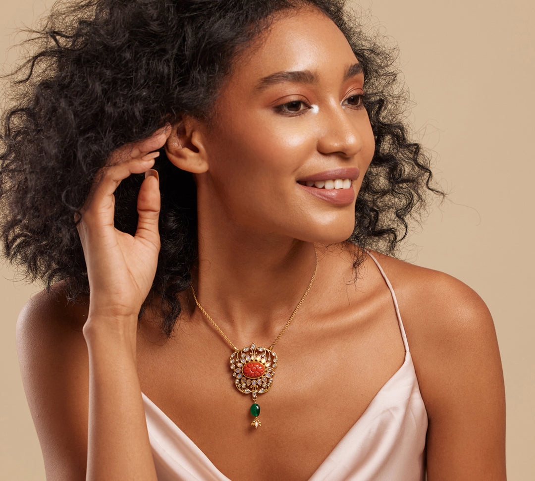 A woman wearing a pink dress showcasing a gold necklace with a large orange semi-precious stone pendant and small green stone accents.
