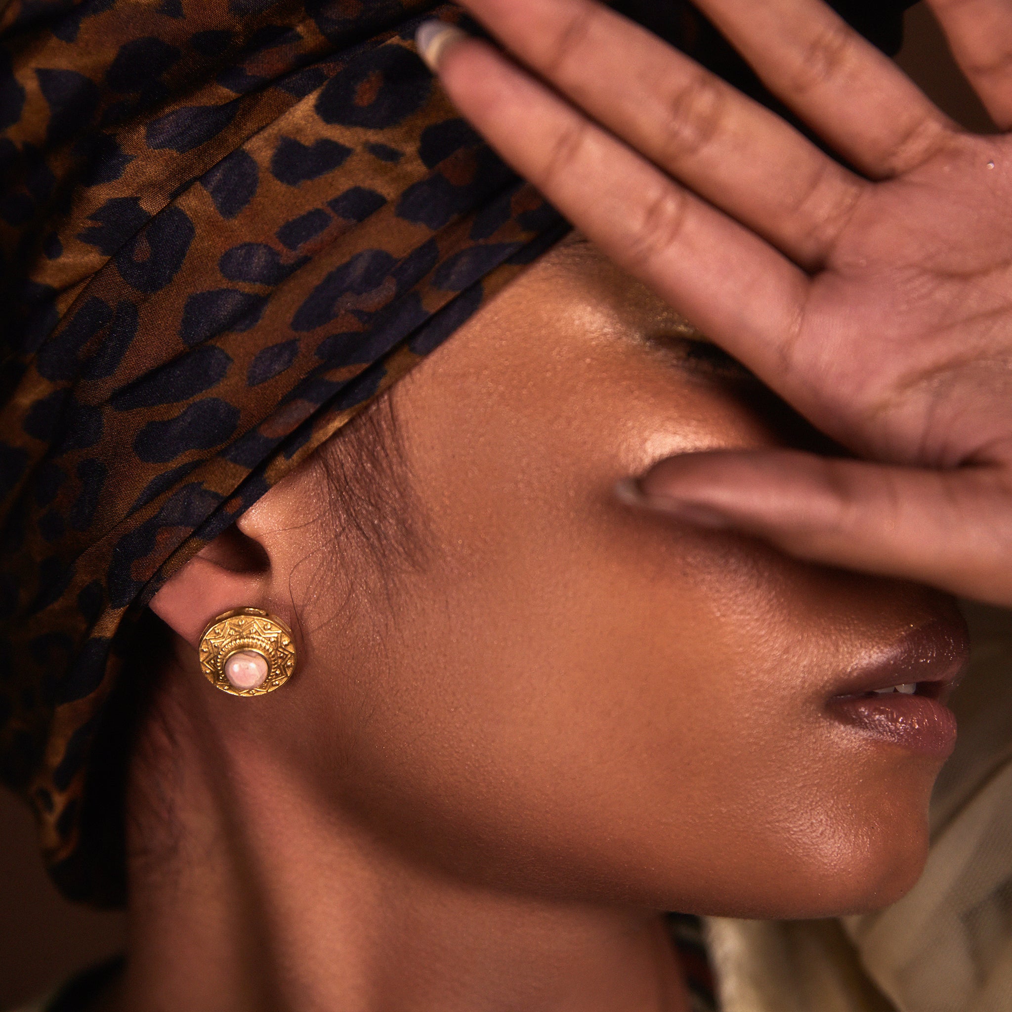 A close-up image of a woman's face, with her hand partially covering her face, highlighting a gold stud earring with a gemstone.