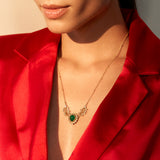 A person wearing a red jacket showcasing a dainty necklace with semi-precious green stones and kundan work.