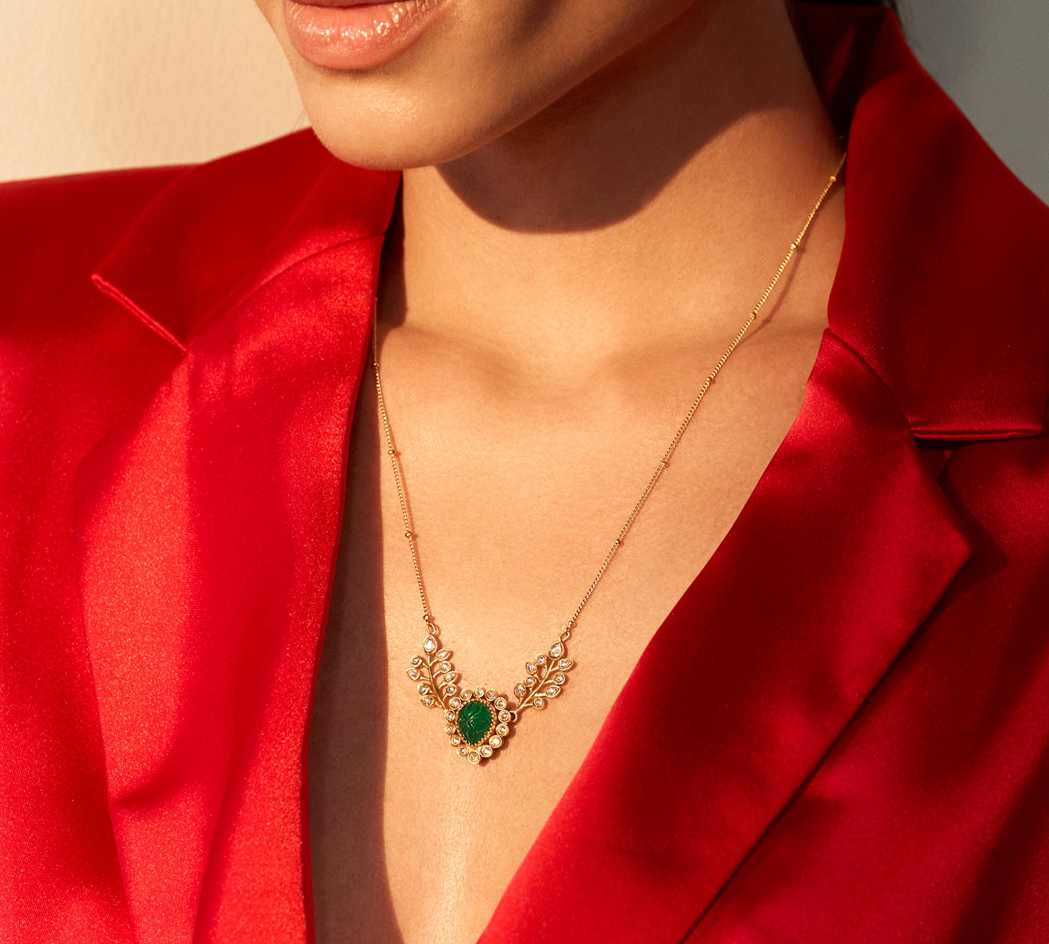 A person wearing a red jacket showcasing a dainty necklace with semi-precious green stones and kundan work.
